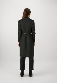 Zwarte, getailleerde lange jas met een riem in de taille, voorzien van een scherpe kraag en een rechte snit, gecombineerd met donkere jeans en zwarte hoge laarzen.