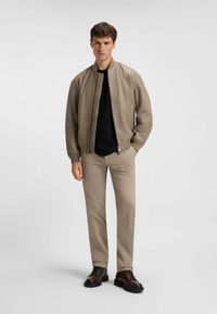 Giacca bomber beige chiaro, camicia nera sotto, pantaloni beige chiaro e stivali marroni con lacci su uno sfondo neutro.
