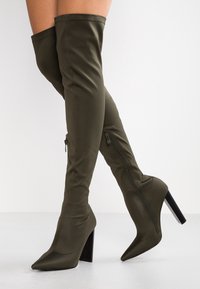 Botas color verde oliva por encima de la rodilla, hechas de un tejido suave, con punta afilada, tacón grueso negro y detalle de cremallera lateral.