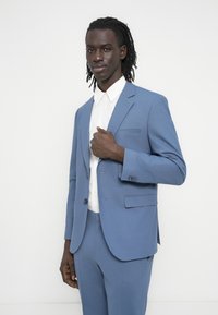 Άνδρας με ντραντλόκς που φοράει ανοιχτό μπλε κοστούμι και λευκό πουκάμισο, κρατώντας το πέτο του σακακιού του μπροστά σε λευκό ομοιόμορφο φόντο.