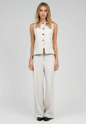 Gilet sans manches beige avec boutons associé à un pantalon large. Tissu lisse avec une coupe ajustée et des lignes nettes. Des chaussures noires complètent le look.