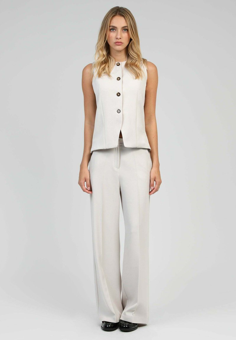 Gilet sans manches beige avec boutons associé à un pantalon large. Tissu lisse avec une coupe ajustée et des lignes nettes. Des chaussures noires complètent le look.