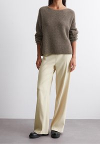 Bruine geribbelde gebreide trui met opgerolde mouwen, gecombineerd met lichtgele flared broek en zwarte plateauzolen, op een neutrale achtergrond.