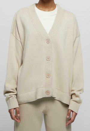 Person trägt eine beige übergroße, geknöpfte Strickjacke über einem weißen Hemd und passenden beigen Hosen, steht vor einem neutralen Hintergrund.