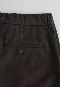 Pantaloni in tessuto marrone con davanti piatto e una tasca sul retro con un bottone nero. La texture appare liscia con un sottile motivo intrecciato.