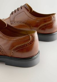 Par de zapatos de vestir de cuero marrón pulido con detalles de brogue y suelas negras sobre una superficie clara.