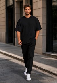 Man walking on city sidewalk wearing black outfit and white sneakers with one hand in pocket and serious expression.