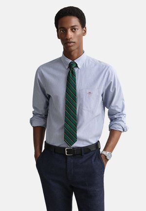 Camisa de vestir a rayas azul claro con cuello abotonado, combinada con una corbata verde con rayas amarillas. Pantalones azul marino y cinturón negro.
