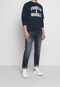 Felpa blu navy con scritta bianca "FRANKLIN & MARSHALL", abbinata a jeans denim scuro strappati e sneakers bianche con dettagli blu navy.