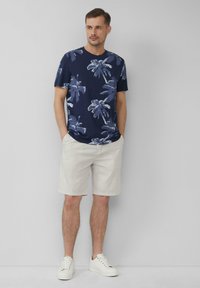 Dunkelblaues T-Shirt mit kurzen Ärmeln und weißem Palmenmuster, kombiniert mit hellbeigen Shorts und weißen Sneakers, vor einem grauen Hintergrund stehend.