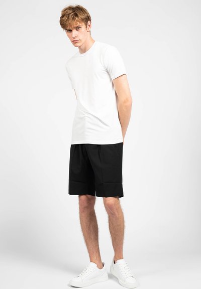 T-shirt bianco in cotone abbinato a pantaloni corti neri, con una vestibilità comoda. Il modello indossa sneaker bianche, in una situazione neutra.