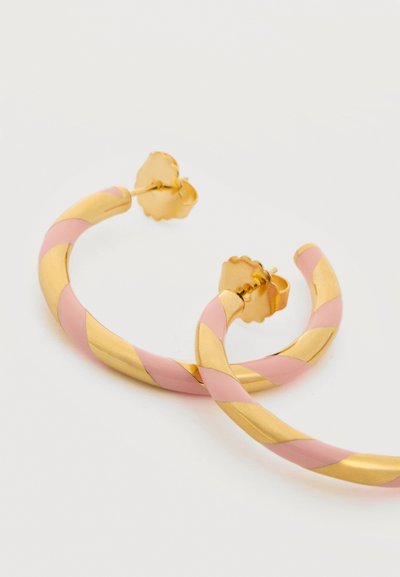 Złote i różowe kolczyki typu koła z paskami i motylkowymi zapięciami na białym tle.