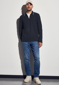 Námořnické polo s dlouhým rukávem a zipovým límcem, s jemně šedými detaily, v kombinaci s světle modrými džíny a bílými teniskami, na jednobarevném pozadí.