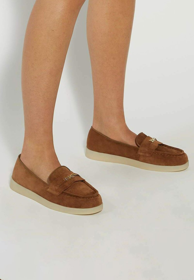 Brown suede loafers featuring a low profile, rounded toe, and a cream-colored sole. Accented with a decorative strap detail.