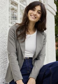 Uśmiechnięta kobieta z długimi ciemnymi włosami, ubrana w szarą marynarkę, białą koszulę i niebieskie dżinsy, siedząca przy białej fakturowanej ścianie i oknie.