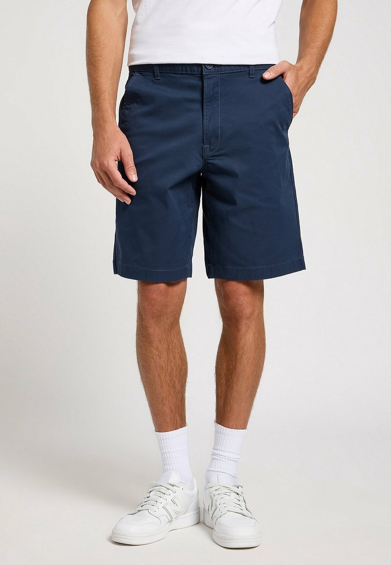 Homme portant un short bleu marine jusqu'aux genoux, des chaussettes blanches, des baskets blanches et une chemise blanche, une main dans la poche.