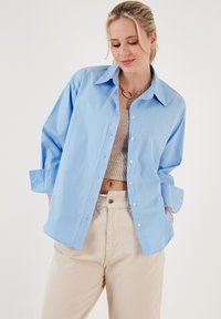 Light blue button-up shirt in cotton blend with a pointed collar, loose fit, and rolled sleeves, worn over a beige ribbed top and beige pants.