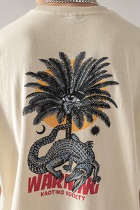 Cremefarbiges T-Shirt mit einem Grafikdesign einer Palme, einem Auge, einem Krokodil und dem Text "WARNUNG" in fettem Rot.