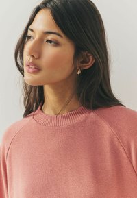 Sweatshirt fuchsia avec un col rond côtelé et des manches raglan, fabriqué en tissu texturé. Le modèle porte une paire de boucles d'oreilles en forme de goutte.