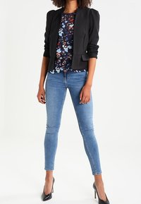 Veste noire à manches bouffantes, blouse fleurie aux tons bleus, jean skinny bleu et escarpins noirs, sur fond blanc.