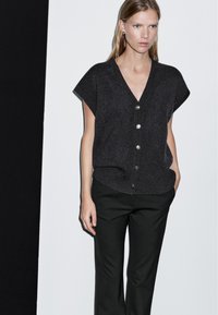 Frau in einem dunkelgrauen ärmellosen Pullover mit Knopfleiste und V-Ausschnitt, kombiniert mit schwarzen Hosen, stehend vor einem schwarz-weißen Hintergrund.