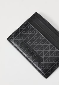 Portefeuille en cuir noir avec un motif géométrique et un logo en relief. Comprend plusieurs emplacements pour cartes et une texture lisse.