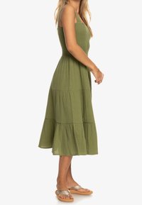 Vestido verde oliva con cuerpo fruncido, falda en capas y tirantes ajustables. Lleva sandalias planas color tostado con detalles metálicos.