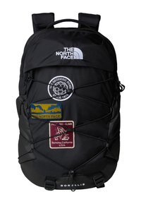 Black backpack with multiple patches, featuring a logo, bungee cord design, and a smooth texture. Includes straps and a zipper closure.