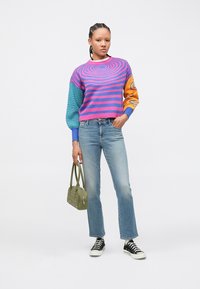 Daudzkrāsains džemperis ar spirālveida un svītrainiem rakstiem, zilās un rozā toņos, kopā ar gaiši zilām džinsu biksēm un melniem sporta apaviem, turot zaļu somu.