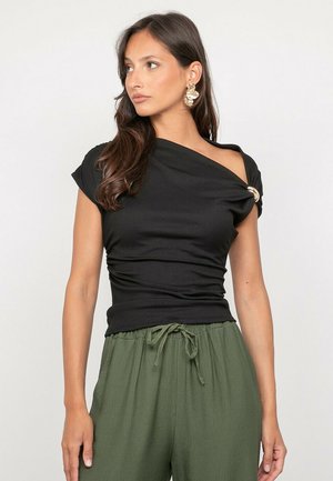 Femme portant un haut asymétrique noir avec boucle dorée et pantalon vert à cordon, regardant sur le côté devant un fond uni.