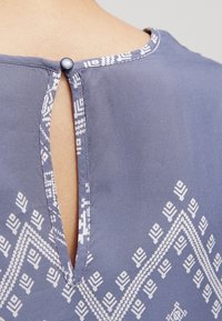 Modrá blůza s geometrickým bílým vzorem, s otvorem ve tvaru klíče na zadní straně, zajištěná modrým knoflíkem, s hladkou texturou látky.