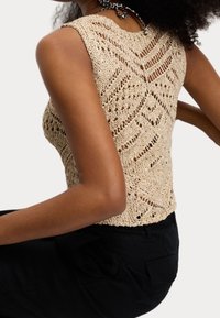 Brede gebreide mouwloze top in beige met een getextureerd ontwerp, gekenmerkt door een open weving en een textuur oppervlak, gecombineerd met zwarte high-waisted broeken.