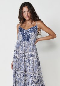 Rochie maxi cu un imprimeu floral albastru și alb, cu bretele subțiri, decolteu în V și un tiv în straturi, realizată dintr-un material ușor.