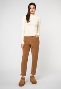 Junge Frau mit langen Haaren, die einen cremefarbenen gerippten Rollkragenpullover, braune hochgekrempelte Cordhosen und braune Plateauschuh-Stiefeletten trägt, steht vor einem weißen Hintergrund.