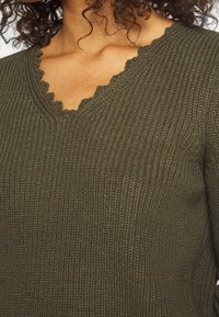 Olivgrüner Strickpullover mit einem gewellten V-Ausschnitt und strukturiertem Stoff. Nahaufnahme des Ausschnitts und der Brustpartie.