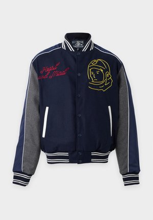Mann trägt eine marineblaue Collegejacke mit Astronauten-Stickerei, weißes Grafik-T-Shirt, beige weit geschnittene Hosen und weiße Sneakers, steht mit den Händen in den Taschen.