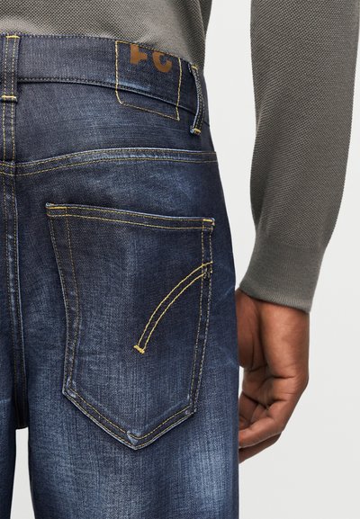 Κοντινό στην πίσω τσέπη ενός μπλε τζιν με κίτρινη ραφή, φορεμένο με γκρι μακρυμάνικο τοπ με υφή, που δείχνει το δεξί χέρι χαλαρό δίπλα στο σώμα.