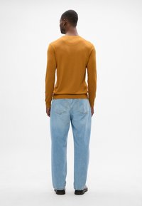 Sweter z dzianiny w kolorze musztardowym z ściągaczami na rękawach, zestawiony z jasnoniebieskimi dżinsami w prostym kroju i czarnymi butami. Neutralne tło podkreśla uwagę na odzieży.