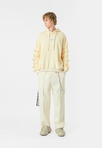 Sudadera amarilla con capucha y mangas de volantes en capas, combinada con pantalones crema claros que tienen un detalle de cadena y zapatos beiges texturizados.