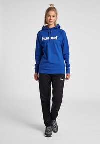 Blauwe hoodie met wit logo, zwarte broek en grijze sneakers. Voorzien van een trekkoordcapuchon, geribde manchetten en een casual pasvorm.