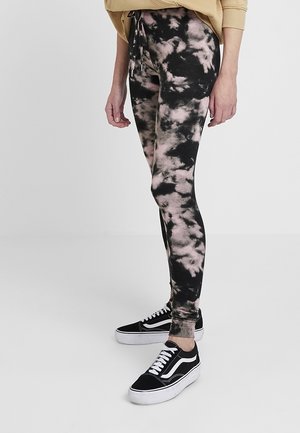 Persoon draagt zwarte en roze tie-dye leggings, een beige trui en zwart-witte veterschoenen, staand op een effen witte vloer.