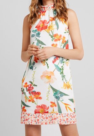 Mouwloze witte jurk met grote oranje, gele en rode bloemmotieven en een kleinere bloemenrand aan de zoom, gedragen door een vrouw met golvend haar.