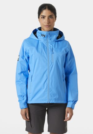 Chaqueta impermeable de color azul claro con capucha, con cierre de cremallera frontal, bolsillos laterales y logo en el pecho. Textura suave, ajuste moderno.