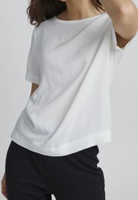 Vit kortärmad topp med en mjuk, slät textur, med rund halsringning och en något lös passform, i kombination med svarta byxor.