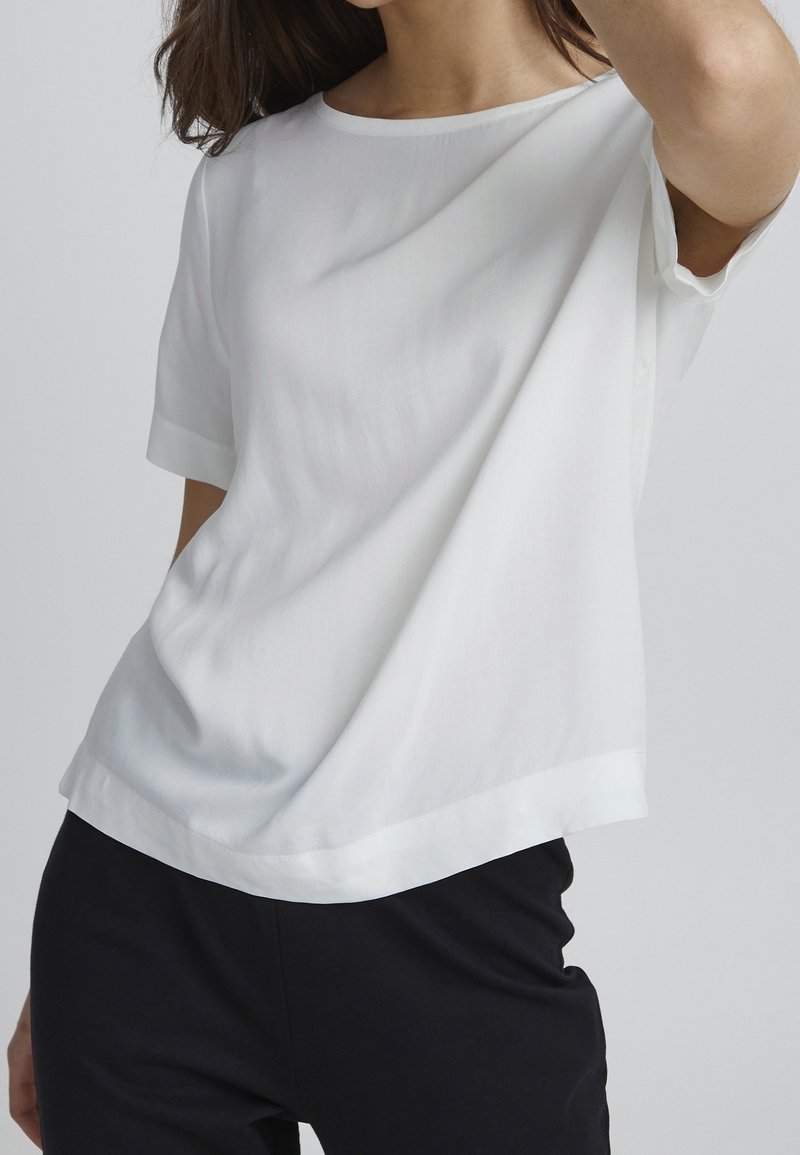 Vit kortärmad topp med en mjuk, slät textur, med rund halsringning och en något lös passform, i kombination med svarta byxor.