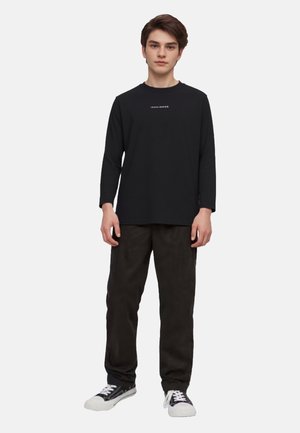 Schwarzes Langarm-T-Shirt mit weißem Text auf der Brust, kombiniert mit dunklen Hosen und schwarzen Sneakern, vor einem schlichten weißen Hintergrund stehend.