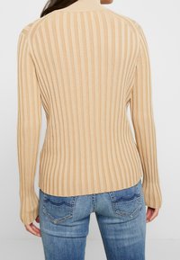 Femme portant un haut beige côtelé à manches longues et un jean en denim bleu, vue de dos sur un fond blanc uni.