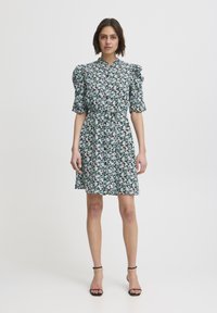 Rochie florală cu mâneci puff scurte, guler cu nasturi, talie strânsă, lungime până la genunchi, bază verde cu flori mici roz și albe. Sandale negre.