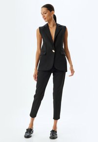 Schwarzer ärmelloser Blazer mit schrägem Revers und einem einzelnen goldenen Knopf, kombiniert mit enganliegenden schwarzen Hosen und glänzenden schwarzen Loafers.