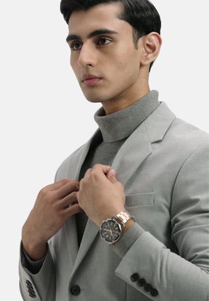 Giacca da completo grigia su un dolcevita grigio, con un orologio in acciaio inossidabile e oro rosa su una mano che aggiusta la giacca.